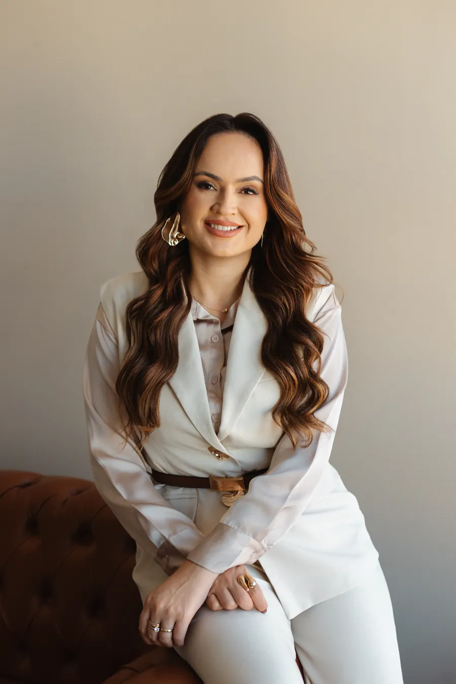 Nathaly de terninho creme, sorrindo, sentada no braço de um sofá de couro.
