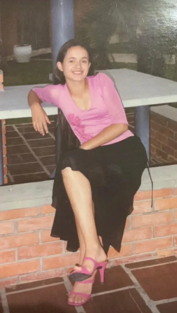 Nathaly muito jovem, sentada em um muro de tijolos, de blusa rosa.